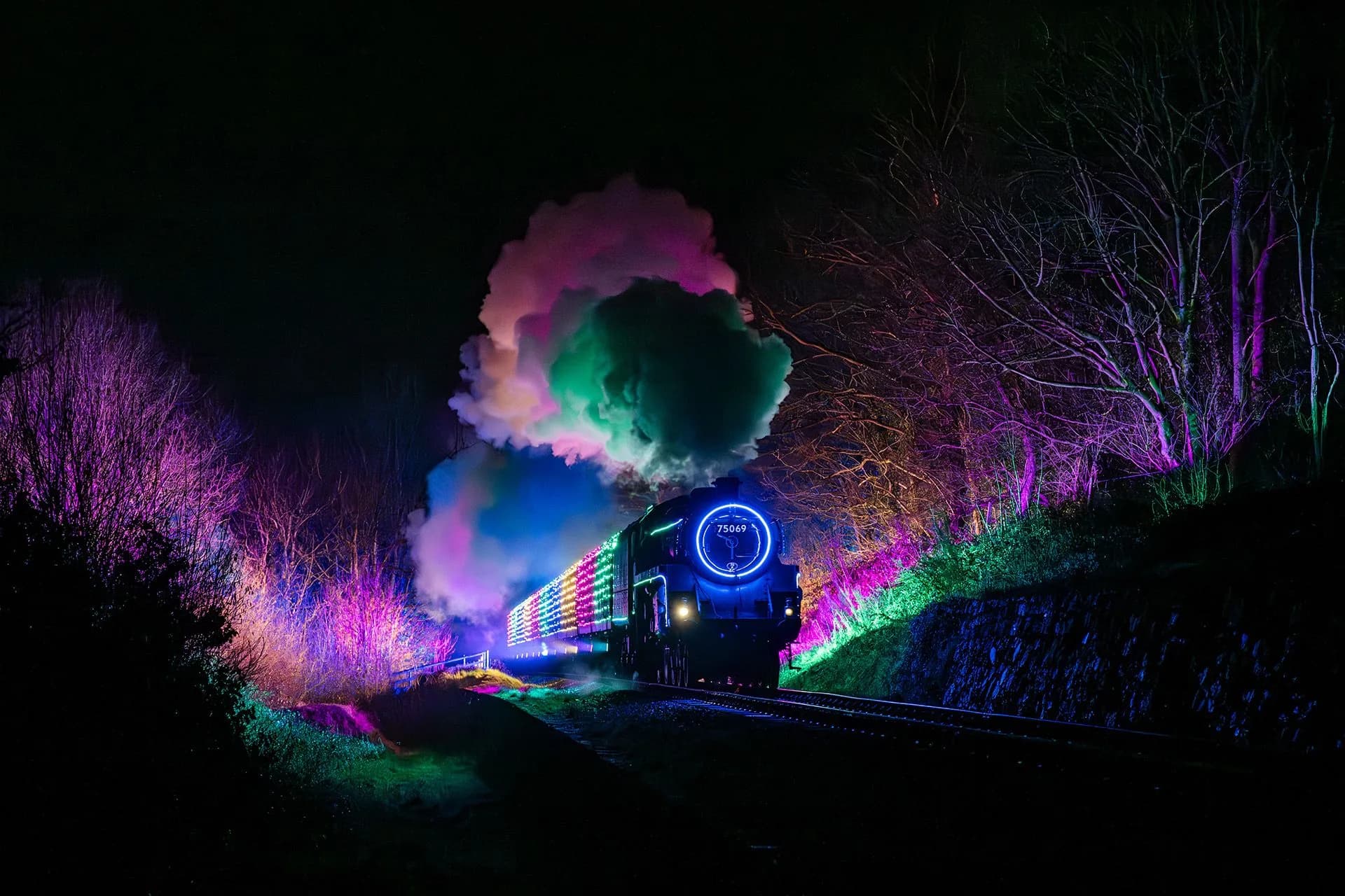 A steam locomotive covered in glowing LED lights travelling down a railway line at night