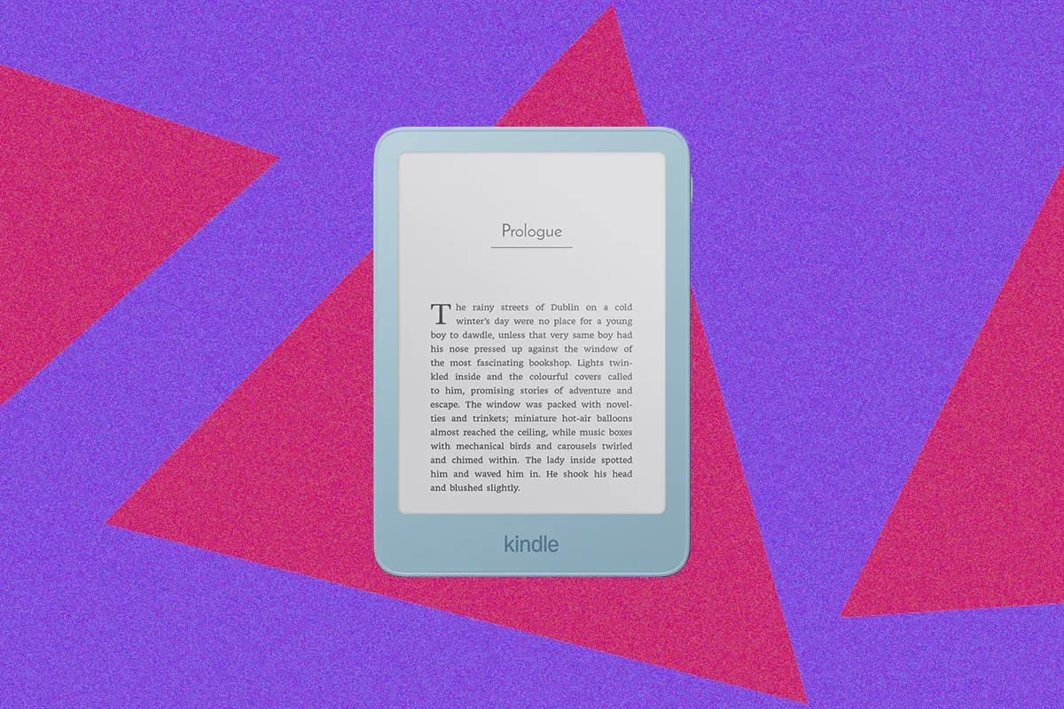 A white Kindle Paperwhite Signature Edition lying flat on a wooden desk, screen off, next to a pair of glasses and an open paperback.
