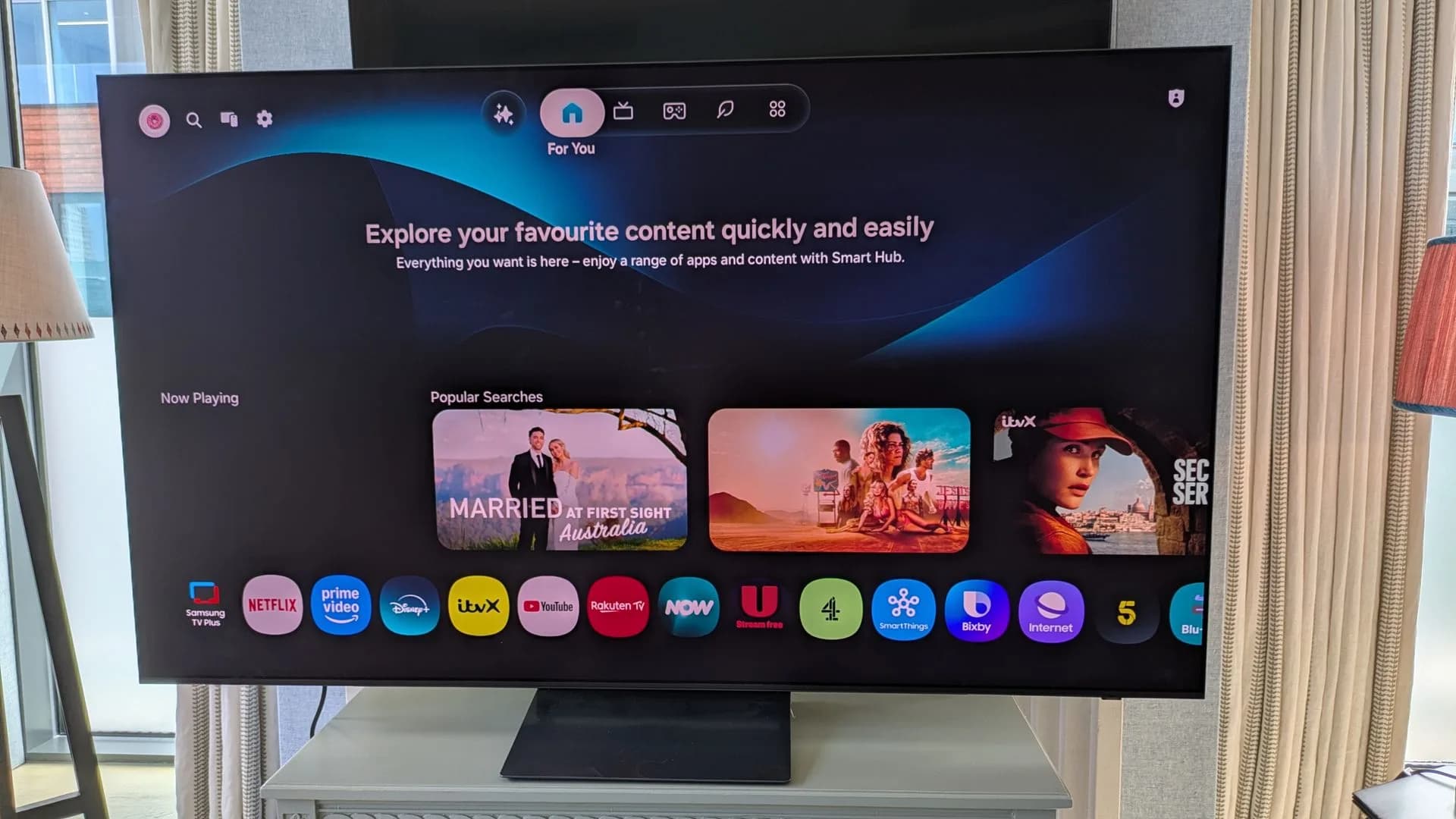 Front view of the Samsung S90H OLED television sitting on a media console in a bright room.