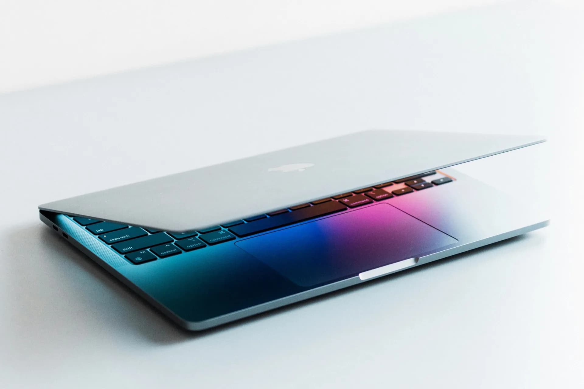 A mockup-style render of a thin, bezel-less MacBook with a glowing OLED display, shown alongside macOS 27.2's new touch-responsive Dock animation.