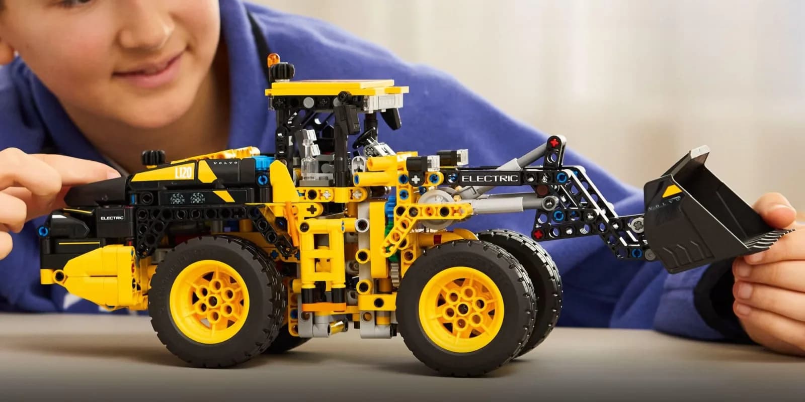 Lego Technic Volvo L120 electric wheel loader model built with yellow beams, black wheels, and a front shovel next to a toy charging station
