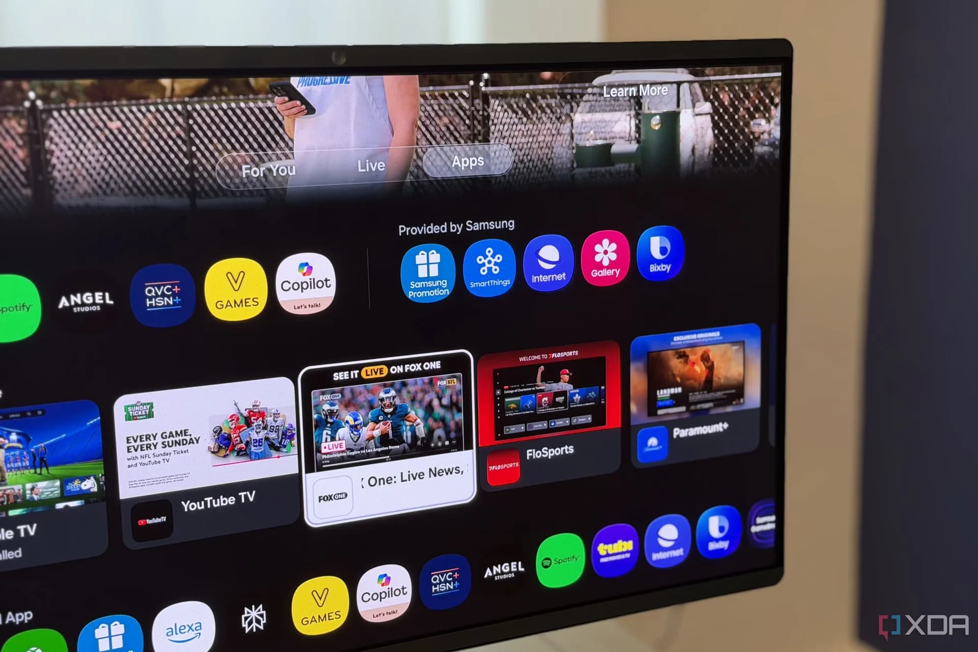 A Samsung smart TV screen displaying a minimal home screen dashboard with app icons and a nature wallpaper, with a smartphone propped up facing the screen.