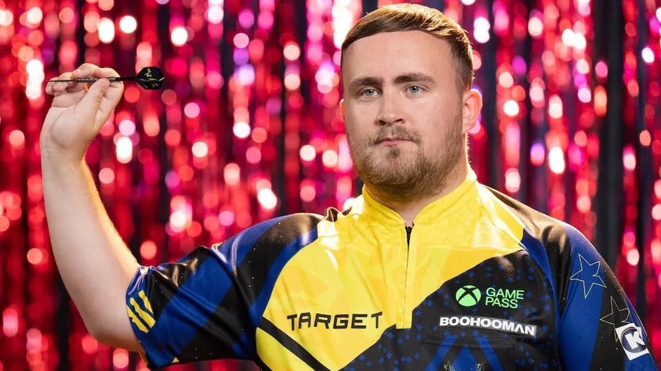 Darts player Luke Littler throwing a dart during a match, wearing a dark playing shirt.